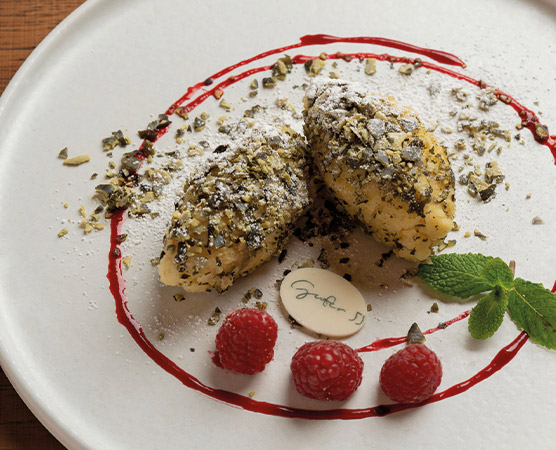 Auf dem Teller befinden sich zwei gefüllte Teigstücke, bestreut mit gehackten Nüssen und Puderzucker. Um sie herum sind Himbeeren und ein spiralförmiger Rand aus roter Sauce. Ein Dekorationsstück mit dem Schriftzug „Gourmet“ ist ebenfalls sichtbar.