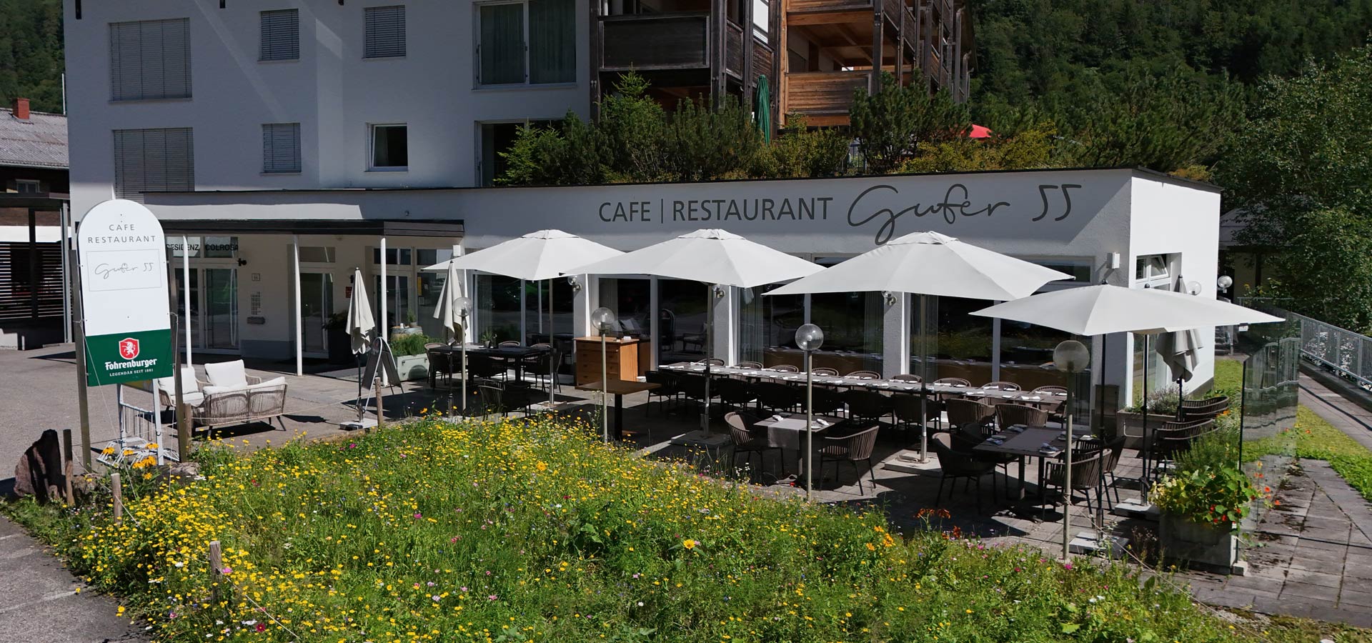 Das Bild zeigt das Café Restaurant Gufer 55 mit einer einladenden Terrasse, die mit Sonnenschirmen und Tischen ausgestattet ist. Vor dem Gebäude wächst eine bunte Blumenwiese. Die Fassade des Restaurants ist modern und freundlich gestaltet.