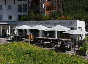 Ein modernes Café und Restaurant mit Außenbereich, ausgestattet mit Sonnenschirmen und Tischen. Der Name "Genuss" ist in stilvoller Schrift an der Wand angebracht. Umgeben von einer blühenden Wiese und einem grünen Hintergrund.