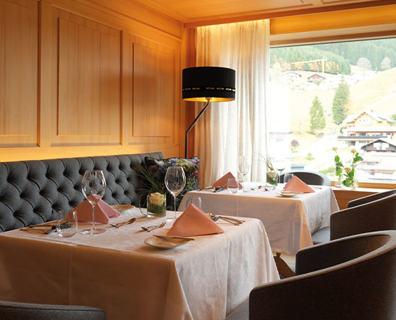 Ein stilvolles Restaurant mit zwei gedeckten Tischen. Jeder Tisch hat eine weiße Tischdecke, Gläser und dekorierte Servietten. Die Wände sind aus Holz, und ein Fenster bietet Blick auf eine grüne Landschaft mit Bergen im Hintergrund. Ein eleganter Lampenschirm beleuchtet den Raum.