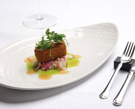 Auf dem Teller befindet sich ein knuspriger Keks, garniert mit frischen Kräutern. Darunter liegt eine bunte Gemüsebeilage aus kleinen, gewürfelten Rüben und einem grünen Dressing. Im Hintergrund ist ein Glas Wein sichtbar.