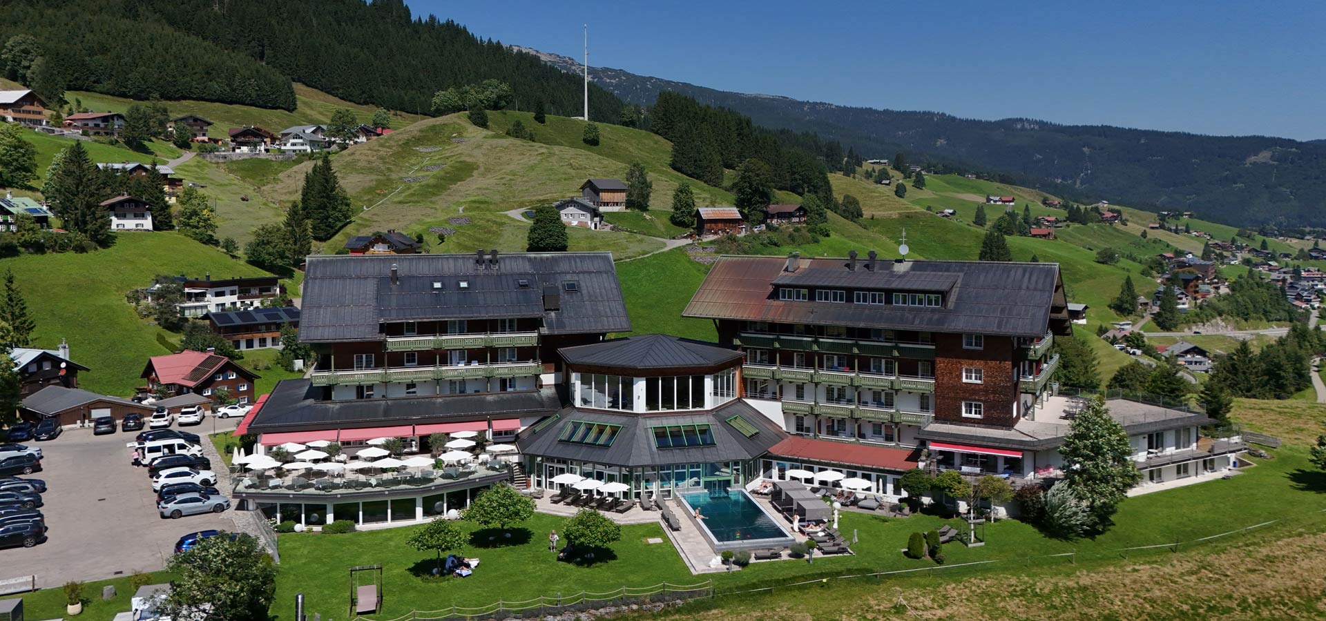 Ein modernes Hotel mit einem Pool liegt in einer malerischen Berglandschaft. Umgeben von grünen Wiesen und Bäumen, sind im Vordergrund Sonnenschirme und Liegen zu sehen. Im Hintergrund erheben sich sanfte Hügel und Wälder.