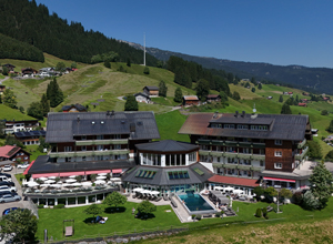 Das Bild zeigt ein Hotelgebäude in einer grünen, hügeligen Landschaft. Es gibt einen Außenpool und Sitzbereiche im Freien. Im Hintergrund sind Bäume und kleine Hütten sichtbar, die eine idyllische Umgebung schaffen. Der Himmel ist klar und sonnig.