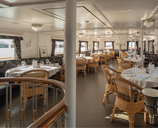 Der Innenraum eines eleganten Restaurants auf einem Schiff, ausgestattet mit runden Tischen und Korbstühlen. Tische sind für Gäste gedeckt, mit weißen Tischdecken und Geschirr. Große Fenster sorgen für eine helle, einladende Atmosphäre. Das Gesamtbild strahlt Komfort und Stil aus.