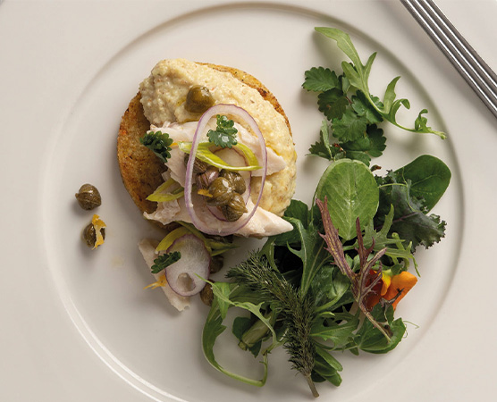 Auf dem Teller befindet sich ein Gericht mit einer Basis aus Brot, belegt mit einer cremigen Sauce, frischem Hühnchen, Kapern und roten Zwiebeln. Daneben liegt ein bunter gemischter Salat mit verschiedenen Blattsalaten und Kräutern.