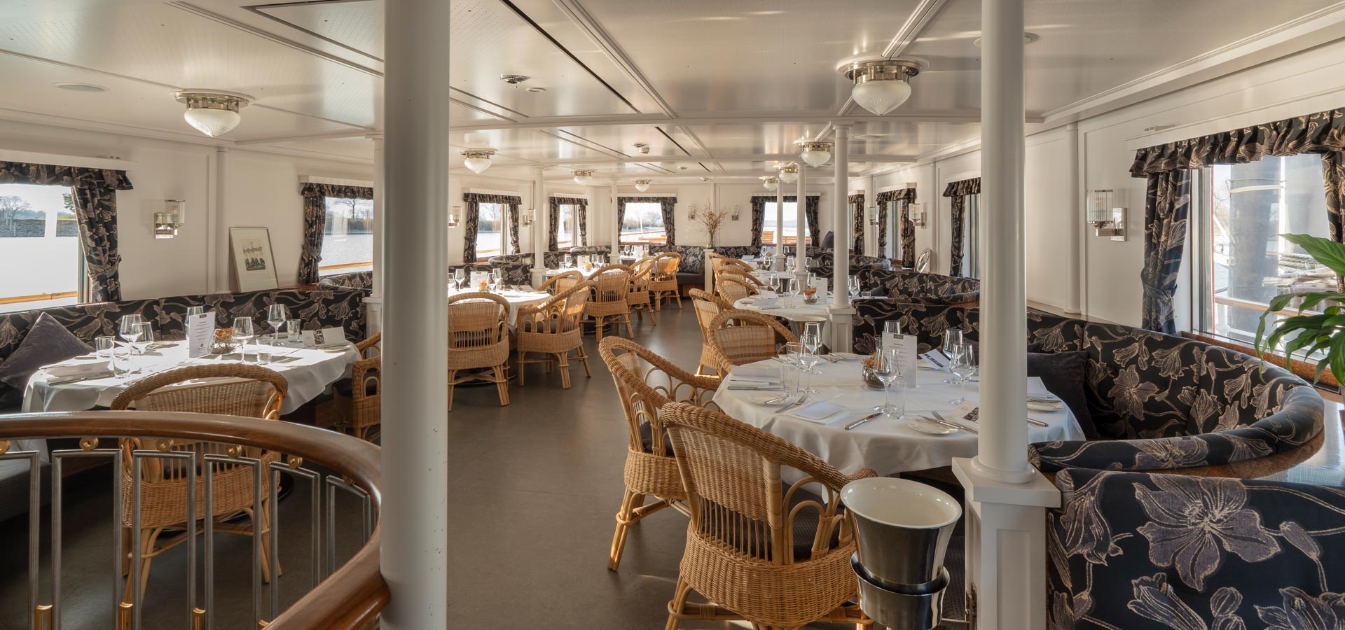Ein elegantes Restaurant auf einem Schiff mit runden Tischen, eingedeckt mit Gläsern und Besteck. Die Wände sind in hellen Farben gehalten und kleine Fenster bieten einen Blick auf das Wasser. Korbsessel und stilvolle Vorhänge verleihen eine gemütliche Atmosphäre.