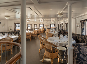 Das Bild zeigt ein elegantes Restaurant im maritimen Stil. Es gibt mehrere gedeckte Tische mit weißen Tischdecken und Stühlen aus Rattan. Große Fenster lassen viel Licht herein und bieten Ausblick auf die Umgebung. Die Wände sind in hellen Tönen gehalten, was eine einladende Atmosphäre schafft.