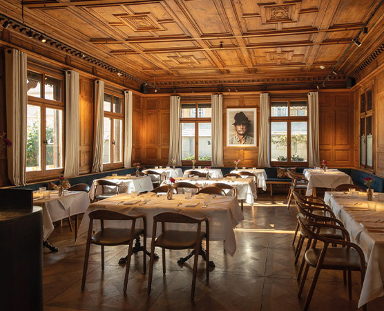 Ein stilvoll eingerichteter Speiseraum mit Holzvertäfelung und großen Fenstern. An den Tischen sind weiße Tischdecken und Geschirr arrangiert. In der Mitte hängt ein Kunstwerk. Der Raum vermittelt eine einladende Atmosphäre für Gäste.