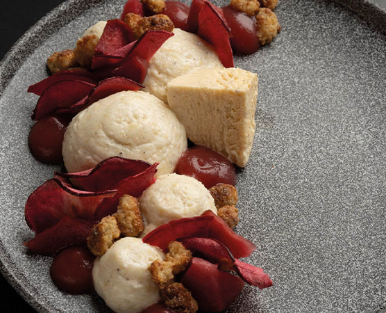 Ein elegantes Dessert auf einem schwarzen Teller, bestehend aus drei runden Portionen einer cremigen Masse, einem stückigen Keks und dekorativen, roten Fruchtpüree- und knusprigen Rosetten aus getrockneten Früchten. Die Anrichtung ist kunstvoll und ansprechend.