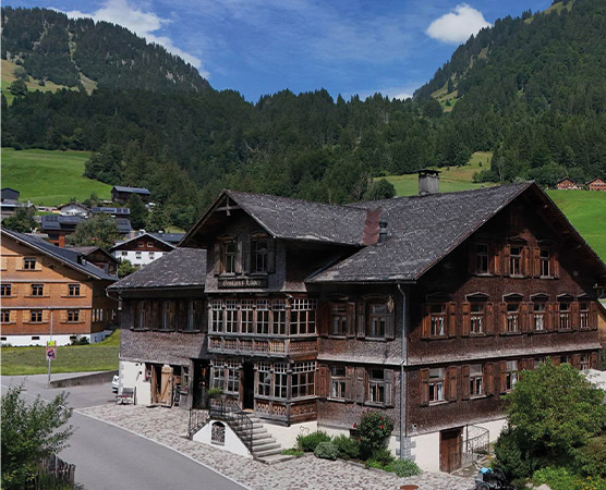 Das Bild zeigt ein traditionelles, mehrstöckiges Holzgebäude im alpinen Stil, umgeben von grünen Bergen. Es handelt sich um ein Gasthaus mit Fenstern im Landhausstil und einer einladenden Veranda. Die Landschaft ist geprägt von sanften Hügeln und weiteren holzverkleideten Gebäuden im Hintergrund.
