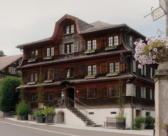 Ein historisches Gebäude mit mehreren Stockwerken und einem charakteristischen roten Holzschnitt. Die Fassade ist mit Fensterläden versehen, und es gibt eine Eingangstreppe. Im Vordergrund stehen Pflanzenkübel und eine Straße ist sichtbar. Das Gebäude strahlt traditionellen Charme aus.