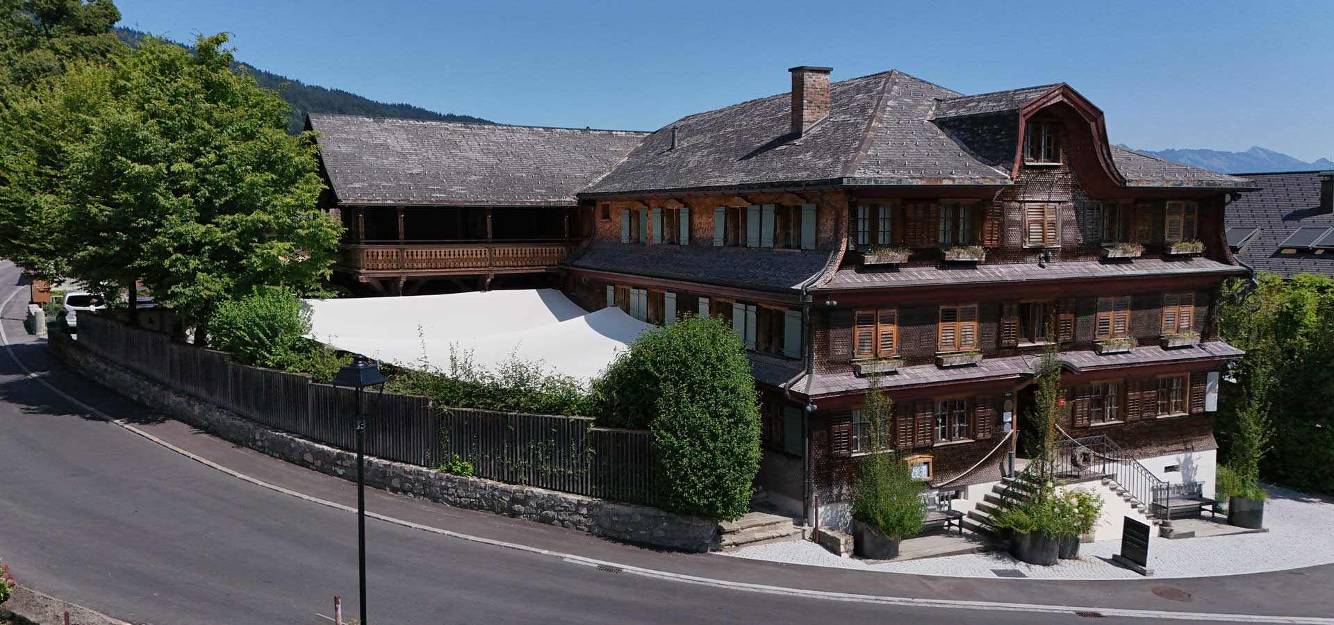 Ein traditionelles Holzgebäude mit mehreren Etagen, großen Fenstern und einem steilen Dach. Umgeben von Bäumen und Pflanzen, steht eine weiße Markise im Vordergrund. Die Straße verläuft halbkreisförmig um das Haus. Im Hintergrund sind sanfte Hügel zu sehen.