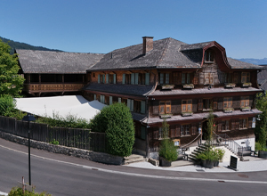 Das Bild zeigt ein traditionelles, mehrstöckiges Holzhaus mit einem gedeckten Eingangsbereich und einer großen Terrasse. Das Gebäude ist von Bäumen umgeben und befindet sich an einer Straße in einer malerischen Umgebung. Der Himmel ist klar und blau.