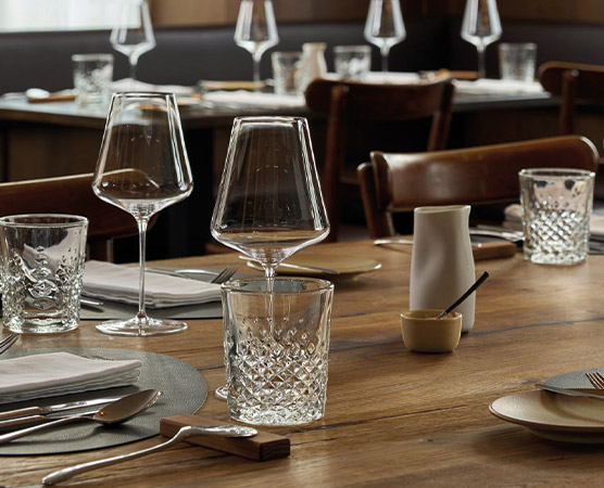 Ein schön gedeckter Tisch in einem Restaurant. Mehrere Gläser, darunter Wassergläser und Weingläser, stehen auf Tischdecken. Es gibt auch eine kleine Flüssigkeitskaraffe und eine Schale mit einem Löffel. Der Tisch ist aus Holz und wirkt einladend.