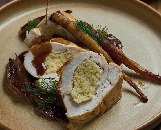 Ein angerichteter Teller zeigt gefülltes Hähnchenfilet, das in dünne Scheiben geschnitten ist. Die Füllung ist sichtbar. Neben dem Hähnchen liegen gebratene Karotten und Zwiebeln, alles leicht mit einer Soße beträufelt. Grünes Dillkraut dekoriert das Gericht.