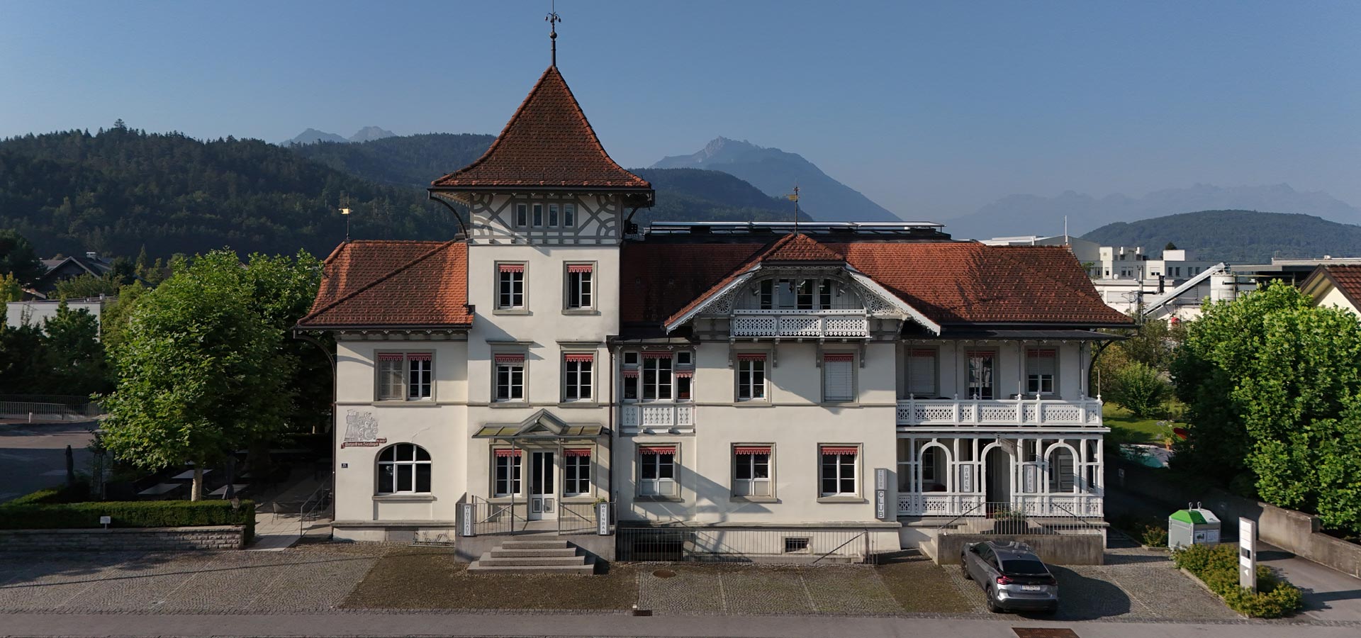 Das Bild zeigt ein historisches Gebäude mit einem markanten, spitzen Dach und einem Erker. Es hat helle Fassaden mit mehreren Fenstern und Verzierungen. Vor dem Gebäude sind Pflanzen und ein Parkplatz zu sehen, vor dem sich ein graues Auto befindet. Im Hintergrund erheben sich Berge.
