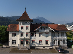 Ein historisches Gebäude mit einem charakteristischen roten Dach und einem Turm. Die Fassade ist hell gestrichen und hat große Fenster sowie einen Balkon. Im Hintergrund sind sanfte Berge und ein klarer Himmel sichtbar.