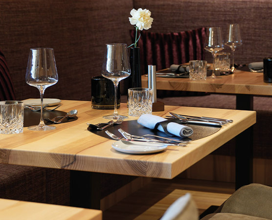 Tischgedeck in einem Restaurant mit eleganten Gläsern, glänzendem Besteck und stilvollen Tellern. Eine weiße Blume in einer kleinen Vase ergänzt die dekadente Atmosphäre. Die Holztische sind einfach, aber modern gestaltet, eingerahmt von weichem, rötlichem Samt.