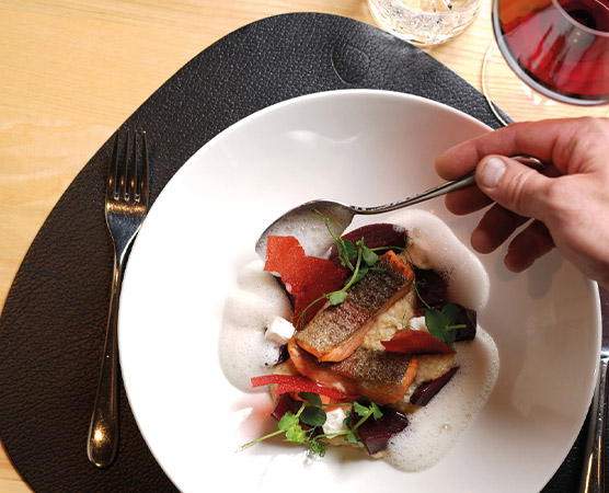 Ein Teller mit gebratenem Fisch, umgeben von buntem Gemüse und einer schaumigen Sauce. Eine Hand hält einen Löffel, während sie gerade das Gericht anhebt. Der Hintergrund zeigt einen Tisch mit Besteck und ein Glas Wein.