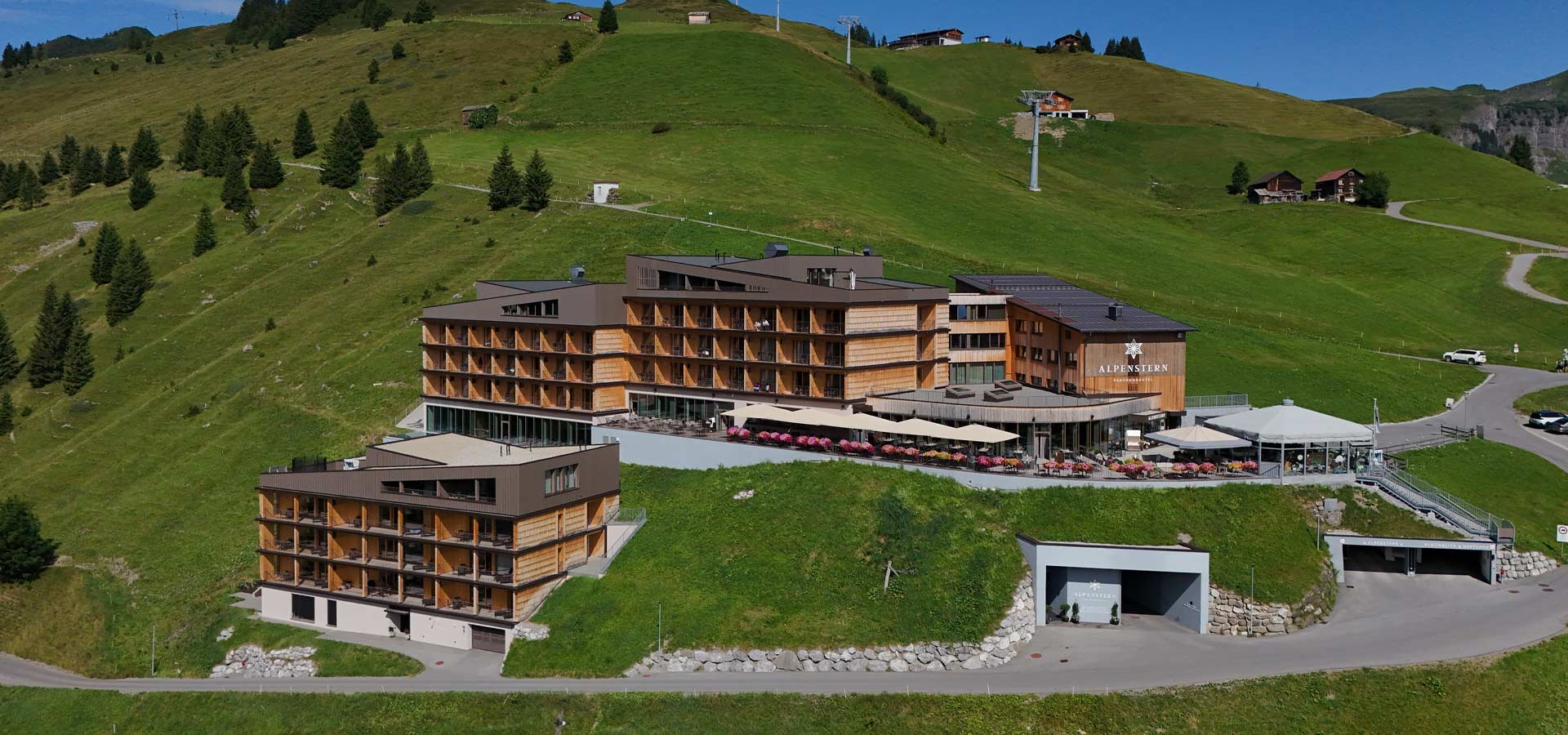 Das Bild zeigt ein modernes Hotel, das in einer bergigen Landschaft liegt. Umgeben von grünen Wiesen und Bäumen, hebt sich das Gebäude mit Holzverkleidung und großen Fenstern hervor. Vor dem Hotel sind Terrassen mit Sonnenschirmen und Blumenarrangements sichtbar.