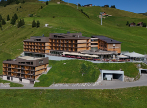 Ein modernes Hotel mit Holzfassade, umgeben von grünen Wiesen und sanften Hügeln. Im Vordergrund sind Terrassen mit Sitzplätzen zu sehen. Im Hintergrund erstrecken sich die Berge, und ein Skilift ist sichtbar. Es vermittelt eine einladende, naturnahe Atmosphäre.