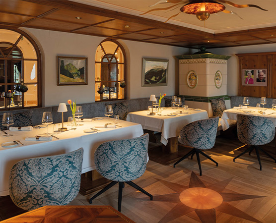 Ein elegantes Restaurant mit Holzvertäfelung und weichen, bläulich gemusterten Stühlen. Tischdecken in Weiß, professionell gedeckte Tische mit Gläsern und Besteck. An den Wänden hängen Bilder. Große Fenster sorgen für eine helle, einladende Atmosphäre.