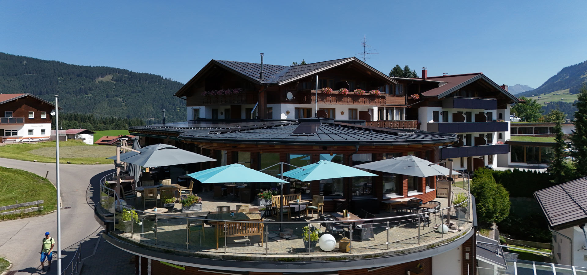 Ein modernes Gebäude mit einem runden, überdachten Außenbereich. Es sind Tische und Stühle unter blauen und grauen Sonnenschirmen aufgestellt. Im Hintergrund sind grüne Hügel und ein blauer Himmel zu sehen, was eine einladende, ruhige Atmosphäre vermittelt.