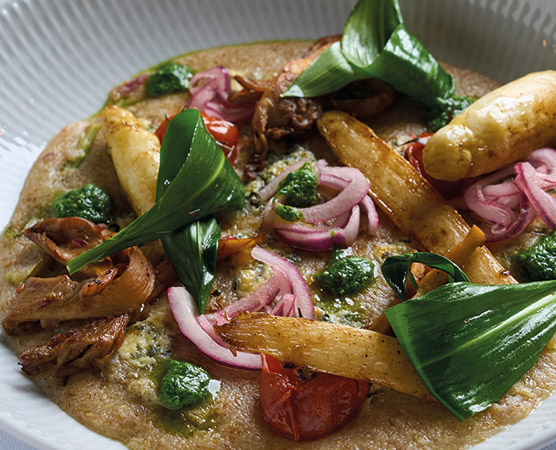 Ein Teller mit einer flachen Brotart, belegt mit gebratenem Hähnchen, knusprigen Kartoffelstäbchen, roten Zwiebeln und frischen Tomaten. Grüner Pesto und Gemüseblätter verleihen dem Gericht einen frischen Look und Geschmack.