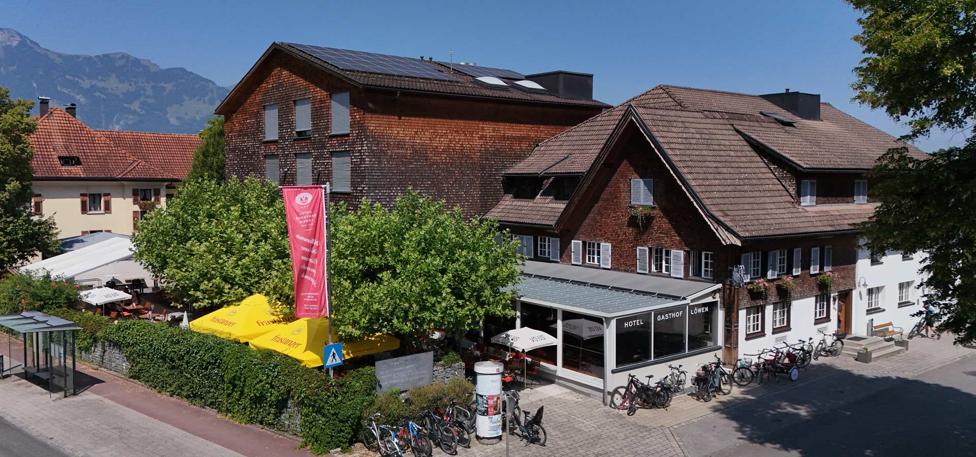 Ein zweigeschossiges Gebäude mit Holzfassade und Satteldach beherbergt das Hotel Gasthof Löwen. Vor dem Gebäude stehen Fahrräder und Tischbereiche im Freien. Bäume und Sonnenschirme verleihen der Umgebung eine einladende Atmosphäre. Im Hintergrund sind Berge sichtbar.