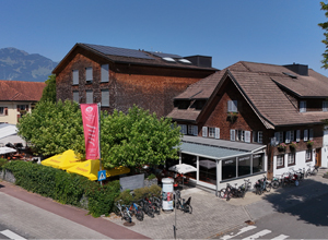 Das Bild zeigt ein ehemaliges, zweigeschossiges Gebäude mit Holzwänden und einem modernen Anbau. Vor dem Gebäude ist ein gemütlicher Außenbereich mit Tischen. Rechts sind Fahrräder geparkt, und ein großes, gelbes Objekt sowie ein rotes Banner sind ebenfalls sichtbar. Im Hintergrund sind Berge zu erkennen.