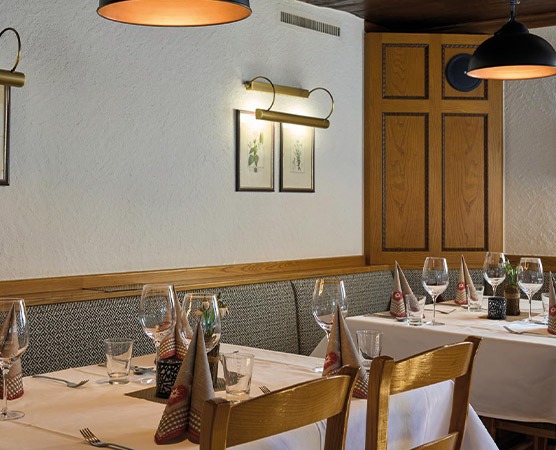 Ein eleganter Restaurantbereich mit gedeckten Tischen. Jeder Tisch hat weiße Tischdecken, Gläser, Besteck und dekorierte Servietten. Die Wände sind mit Pflanzenbildern und Holzvertäfelungen geschmückt. Warme Lichtquellen schaffen eine einladende Atmosphäre.