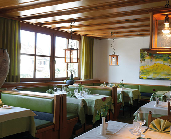 Ein einladendes Restaurant mit holzverkleidetem Decken-Design. Die Tische sind mit grünen Tischdecken und Rosen dekoriert. Große Fenster lassen Tageslicht herein und bieten einen Blick nach draußen. An den Wänden hängen stilvolle Gemälde. Der Raum strahlt eine warme, gemütliche Atmosphäre aus.