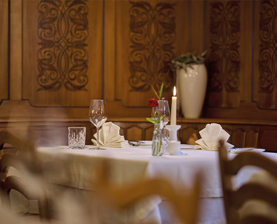 Ein elegant gedeckter Tisch in einem Restaurant, umgeben von Holzvertäfelungen. Auf dem Tisch stehen Weingläser, ein Wasser- und ein Kerzenhalter. In einer kleinen Vase befindet sich eine Blume. Die Tischdekoration ist schlicht und einladend, mit gefalteten Servietten.