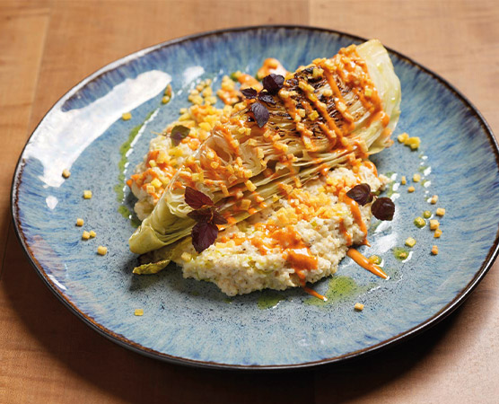 Das Bild zeigt einen Teller mit gegrilltem Gemüse, speziell zwei Hälften von Romanasalat, auf einer cremigen Beilage aus Couscous. Der Salat ist mit einer orangenen Sauce und kleinen Kräuterblättern garniert, was das Gericht ansprechend präsentiert.