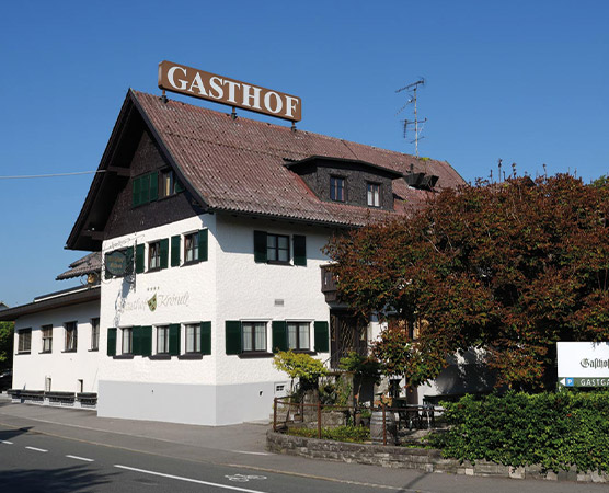 Das Bild zeigt einen Gasthof mit einem typisch bayerischen, ländlichen Stil. Das Gebäude ist zweigeschossig, hat ein Satteldach und ist in hellen Farben gehalten. Die Fenster sind grün gestrichen. Oben prangt das Schild mit der Aufschrift "GASTHOF". Um das Gebäude sind Bäume und Pflanzen angeordnet.