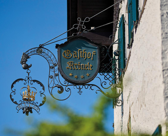 Das Bild zeigt ein schmiedeeisernes Schild mit der Aufschrift "Gasthof Köniele" in goldener Schrift. Das Schild ist an einem Gebäude angebracht und hat eine elegante, geschwungene Form, die durch einen kleinen Kronenanhänger ergänzt wird. Der Himmel im Hintergrund ist blau.