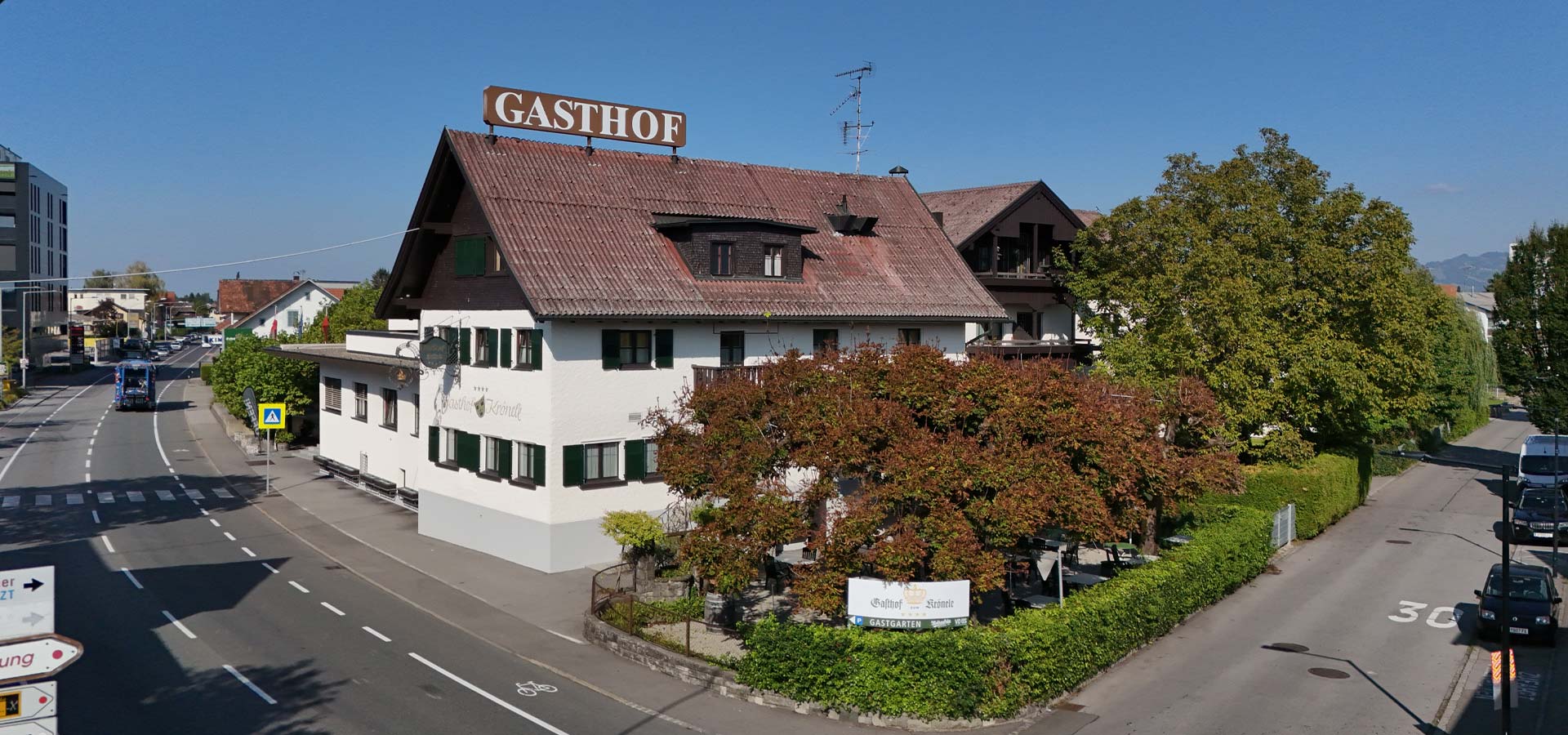 Ein Gasthof mit dem Schild „Gasthof“ auf dem Dach. Das Gebäude hat weiße Wände und grüne Fenster, umgeben von Bäumen und Sträuchern. Im Vordergrund ist die Straße zu sehen, die am Gasthof vorbeiführt. Der Himmel ist klar und blau.