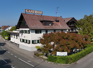 Ein traditioneller Gasthof mit einem großen, verzierten Schild mit der Aufschrift "GASTHOF" auf dem Dach. Das Gebäude hat grüne Fensterläden und ist von Bäumen umgeben. Es befindet sich an einer Straßenkreuzung in einer ländlichen Umgebung.