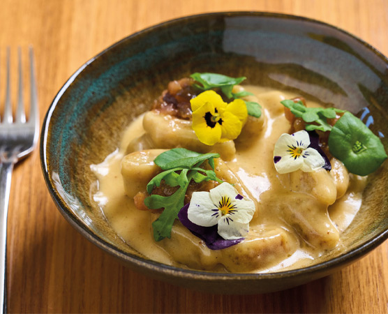 Ein Teller mit einer bunten Mischung aus gegrilltem Fleisch, gebratenen Kartoffeln, roten Zwiebeln und frischen Kräutern. Die Speisen sind ansprechend angeordnet und werden von grünen Sauce und buntem Gemüse begleitet.