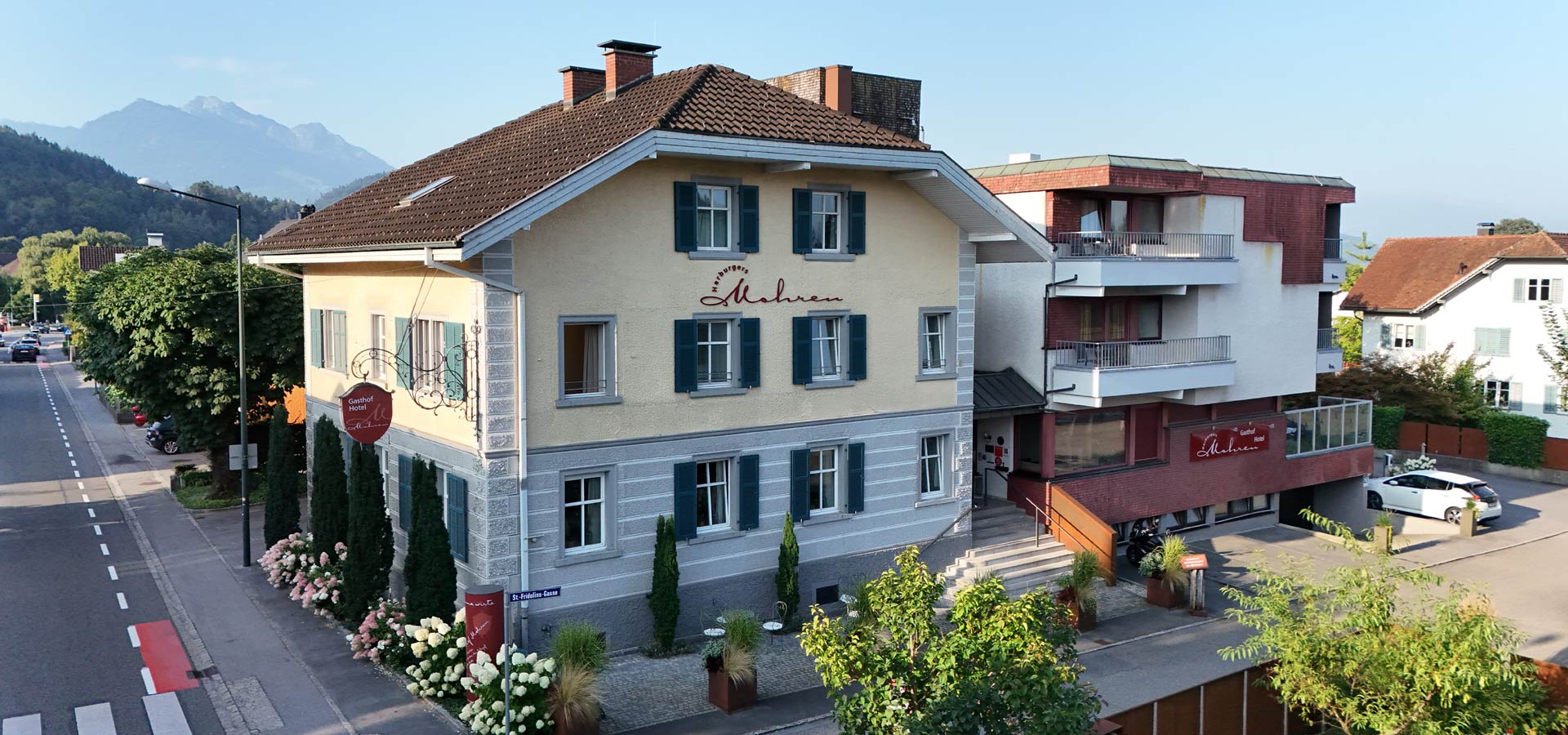 Eine bildschöne Gasthausansicht mit blauen Fensterläden und einem einladenden Eingangsbereich. Im Vordergrund stehen blühende Sträucher. Hinter dem Gasthaus sind majestätische Berge zu sehen. Der Himmel ist klar und die gesamte Szene vermittelt eine ruhige, einladende Atmosphäre.