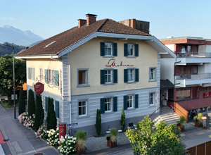 Das Bild zeigt ein zweistöckiges, gelbes Gebäude mit grünen Fensterläden. Der Schriftzug "Hotel" ist sichtbar. Vor dem Haus befinden sich einige Sträucher und Pflanzen. Im Hintergrund sind Berge zu erkennen, was auf eine landschaftlich reizvolle Lage hinweist.