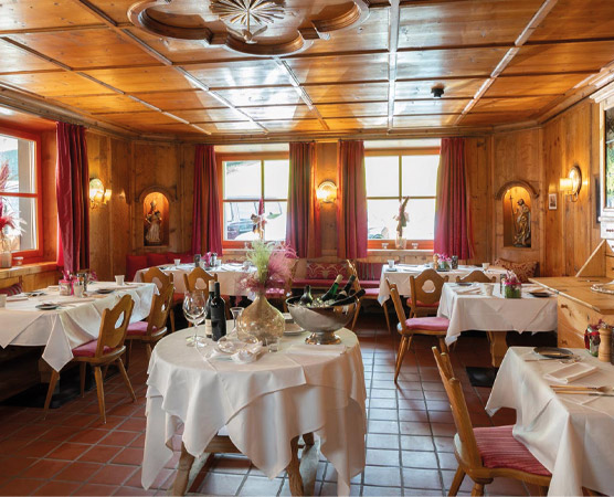 Ein gemütliches Restaurant mit Holzvertäfelung und roten Vorhängen. Mehrere Tische sind mit weißen Tischdecken eingedeckt, dekoriert mit Gläsern und Besteck. Fenster lassen Tageslicht herein, und auf den Wänden sind kleine Bilder zu sehen. Der Raum vermittelt eine einladende Atmosphäre.