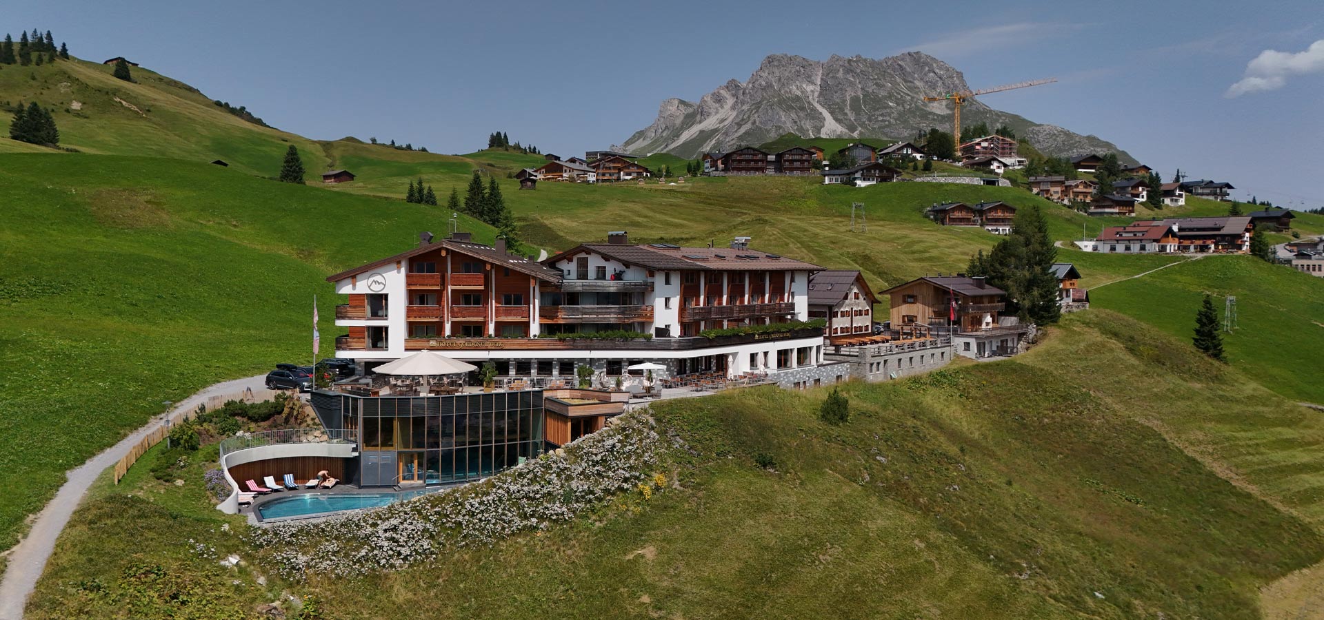 Ein modernes Hotel in einer hügeligen Alpenlandschaft. Im Vordergrund ist ein Poolbereich sichtbar, umgeben von Blumen. Im Hintergrund sind weitere alpine Hütten und Berge, die eine malerische Kulisse bieten. Der Himmel ist klar und die Umgebung wirkt einladend und naturnah.