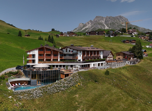 Das Bild zeigt ein charmantes Hotel in einer hügeligen Landschaft, umgeben von grünen Wiesen und Bergen. Das Gebäude mit Holzfassade verfügt über mehrere Etagen und einen modernen Anbau mit einem Poolbereich. Im Hintergrund sind weitere Häuser und hohe Berge sichtbar.