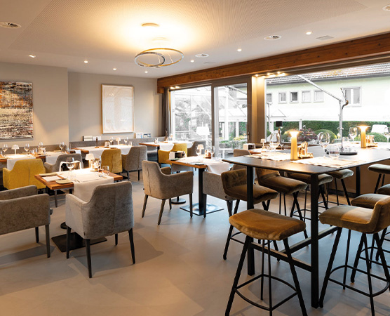 Ein modernes Restaurant mit eleganter Einrichtung. Es gibt runde und hohe Tische mit Stühlen. Die Atmosphäre ist einladend und beleuchtet durch warmes Licht. Fenster zeigen einen Blick nach draußen. Der Raum wirkt sauber und stilvoll, ideal für gehobenes Essen.
