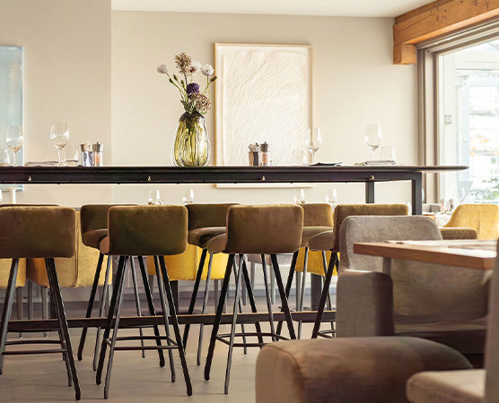 Ein heller Innenraum eines Restaurants mit einer langen Theke, ausgestattet mit stilvollen Barhockern. Auf der Theke steht eine Vase mit bunten Blumen, und es sind Gläser und Besteck sichtbar. Der Raum vermittelt eine einladende und moderne Atmosphäre.