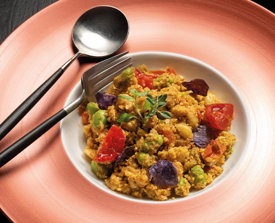 Eine weiße Schüssel mit buntem Gemüse und gekochtem Getreide ist auf einem rosa Teller angerichtet. Oben auf der Mischung befindet sich ein Zweig frischer Oregano. Neben der Schüssel liegen ein Löffel und eine Gabel aus Edelstahl.