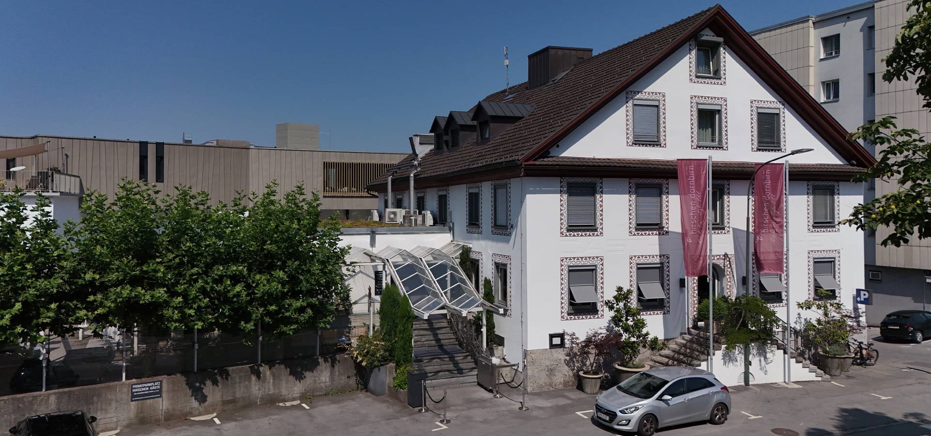 Ein helles, dreigeschossiges Gebäude mit einem giebelbegrünten Dach und dekorativen Fassadenelementen. Vor dem Eingang befindet sich eine Treppe mit einem Glasdach. Um das Gebäude sind Bäume und parkende Autos zu sehen. Im Hintergrund steht ein moderner, schlicht gestalteter Gebäudekomplex.