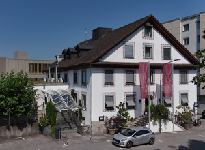 Ein historisches, dreistöckiges Gebäude mit einem Satteldach und weißen Wänden. Vor dem Eingang befinden sich zwei große rosa Banner. Eine Rampe führt zum Eingang, um den Zugang zu erleichtern. Im Vordergrund steht ein silbernes Auto.