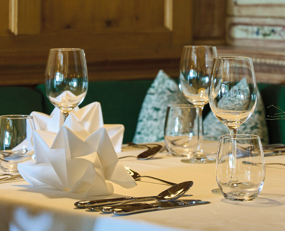 Ein elegant gedeckter Tisch mit mehreren Gläsern, Besteck und einer kunstvoll gefalteten Serviette aus weißem Papier. Im Hintergrund sind grüne Kissen zu sehen. Der Tisch vermittelt eine festliche Atmosphäre.